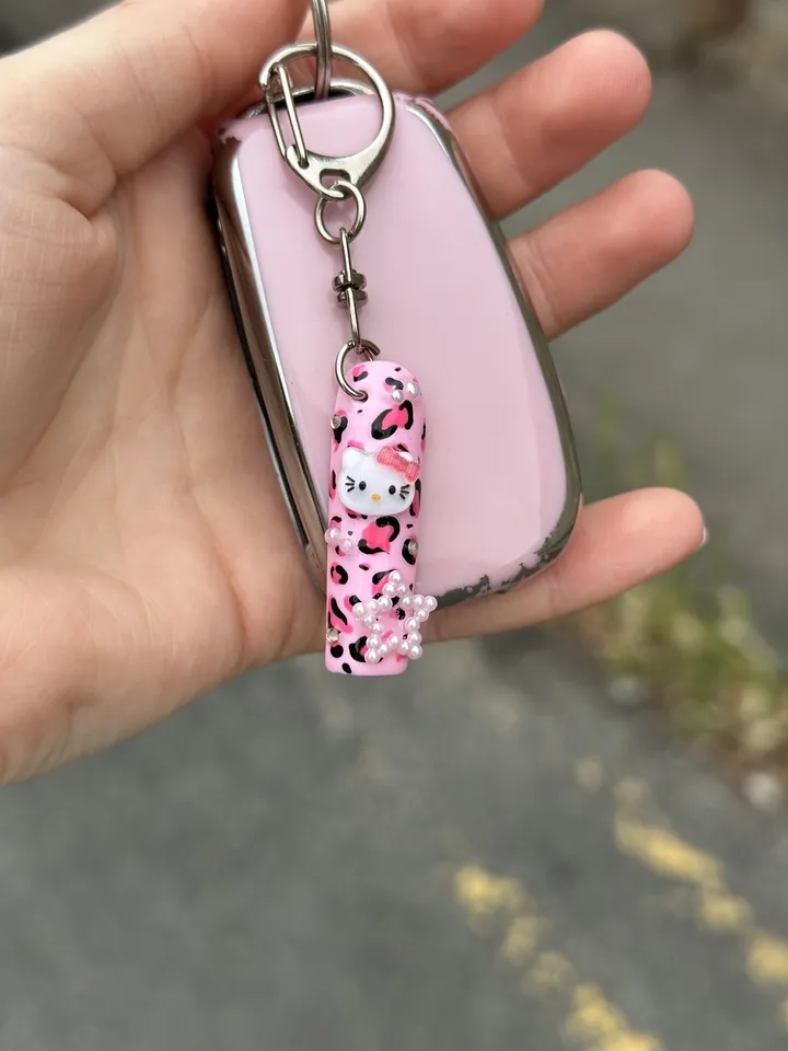 A pink leopard nail charm with a cartoon cat and pearls, hanging from a car key